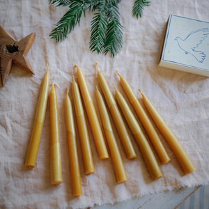 Può includere: Una collezione di candele affusolate giallo dorato disposte su un panno rosa chiaro. Sono presenti anche un rametto di aghi di pino verdi e una piccola scatola con un'illustrazione di una colomba. Una stella di legno aggiunge un tocco rustico.