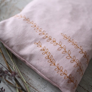 Peut inclure: Une pochette en tissu rose avec un motif brodé décoratif de feuilles brunes. La pochette est posée sur une surface en bois clair.