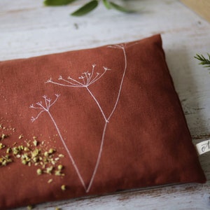 Può includere: Un cuscino termico in lino color ruggine con un motivo floreale ricamato bianco. Il cuscino è riempito di erbe essiccate e ha un'etichetta "cotone" attaccata.