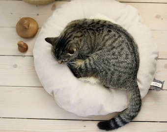 May include: A gray tabby cat curled up in a beige round cat bed with a gray label that says "Olli" on the side.