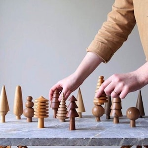 Könnte beinhalten: Ein Set aus Holzspielzeugbäumen in verschiedenen Formen und Größen, darunter Kegel, Zylinder und gestapelte Ringe. Die Bäume sind auf einer hellgrauen Oberfläche angeordnet, wobei eine Hand hineinreicht, um einen der Bäume zu verschieben.
