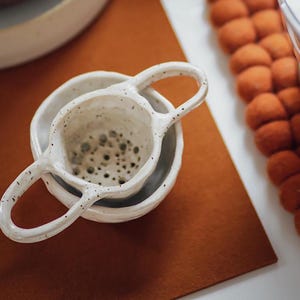 May include: White ceramic tea strainer with two handles. The strainer is nested inside a white ceramic bowl.