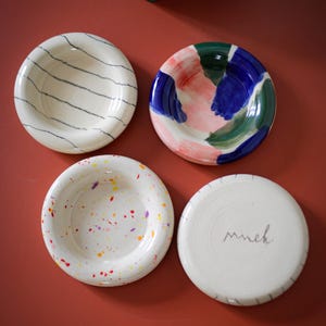 Op de afbeelding: Vier kleine, witte keramische kommen met kleurrijke ontwerpen. Eén kom heeft blauwe strepen, een andere roze, blauwe en groene penseelstreken, een derde heeft kleurrijke verfspatten en de laatste heeft het woord "much".