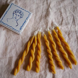 Può includere: Sei candele di cera d'api gialle intrecciate su un tessuto di lino marrone chiaro. Le candele sono disposte in fila, con le punte delle candele rivolte verso il lato destro dell'immagine. Una piccola scatola blu e bianca con un disegno di una testa umana si trova nell'angolo in alto a sinistra dell'immagine.