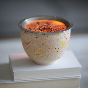 Op de afbeelding: Een kleine, witte keramische kom met een gespikkeld patroon in tinten geel, roze en rood. De kom is gevuld met een rood, oranje en bruin mengsel en staat op een stapel boeken.