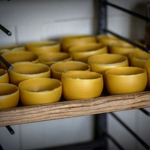 Può includere: Primo piano di uno scaffale di legno pieno di file di candele di cera d'api gialle. Le candele sono di diverse dimensioni e forme, e sono tutte disposte ordinatamente sullo scaffale.