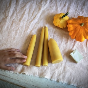 Può includere: Quattro candele di cera d'api gialle e due zucche gialle su una tovaglia di lino bianco.