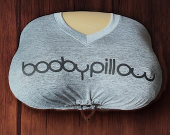 May include: A gray V-neck pillow with the text "booby pillow" printed in black. The pillow is designed to resemble a torso and is set against a brown wooden background.