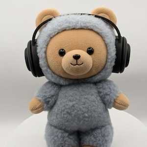 May include: A plush teddy bear wearing a gray hooded outfit and black headphones. The bear has a tan face, ears, and paws, with black eyes and a stitched smile. The bear is standing on a white surface.