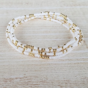 May include: A white and gold beaded bracelet with multiple strands of tiny beads. The bracelet is made with white seed beads and gold accent beads.