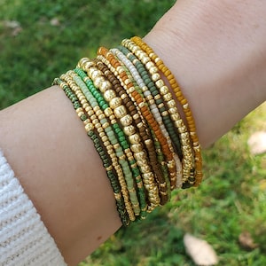 May include: A collection of beaded bracelets in shades of green, gold, and brown. The bracelets are made of small, round beads and larger, gold-colored beads. The bracelets are stacked on a wrist, with a blurred green background.