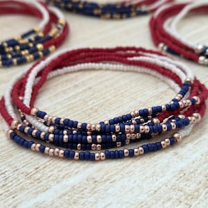 May include: Stackable beaded bracelets in red, white, and navy blue. The bracelets feature small, colorful beads strung on elastic cords. Some bracelets have gold-colored beads interspersed with the blue beads. The bracelets are arranged on a light-colored wooden surface.