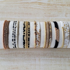 May include: A set of eight beaded bracelets in various colors and patterns, including black, white, gold, and copper. The bracelets are stacked together on a white surface.