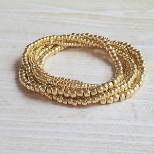 May include: A gold beaded bracelet made of multiple strands of small, round beads. The bracelet is coiled into a circle and is lying on a light wood surface.