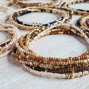 May include: A close-up of a multi-layered beaded bracelet with brown, gold, and cream colored beads. The bracelet is made of small, round beads and has a delicate, feminine look.