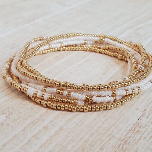 May include: A multi-strand bracelet made of small gold and white beads. The bracelet is layered and creates a delicate and elegant look.