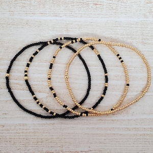 May include: Four delicate bracelets made of black and gold beads. The bracelets are stacked on top of each other, creating a layered look.