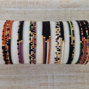 May include: A collection of seven multi-strand beaded bracelets featuring small glass seed beads in various color combinations including black, orange, green, purple, white, and metallic gold accents arranged in repeating patterns and solid rows.