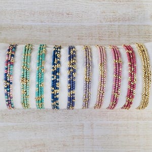 May include: A set of ten beaded bracelets in various colors, including pink, blue, green, and gold. The bracelets are arranged in a row on a white surface.