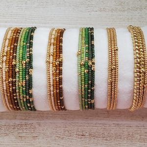 May include: An assortment of beaded bracelets in various colors, including gold, green, brown, and white. The bracelets are arranged side by side on a white surface, showcasing their different designs and textures. The bracelets are made of small beads.