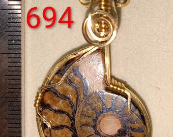 May include: A pendant featuring a fossilized ammonite encased in a gold-colored wire frame. The fossil displays intricate patterns in shades of brown, blue, and beige. The number "694" is visible in red. The pendant has a loop for a chain.