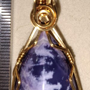 May include: A gold-colored pendant with a triangular, purple and white stone. The stone is encased in a gold wire frame. The pendant has a loop at the top for a chain. The number "777" is written in red.