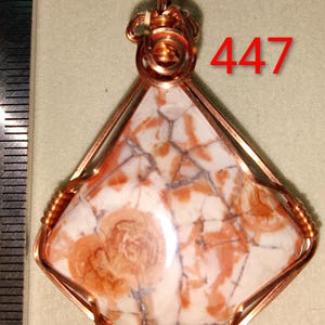 May include: A handcrafted pendant featuring a unique, patterned stone in shades of white and orange, encased in copper wire. The pendant has a triangular shape with a beaded border and the number "447" in red.