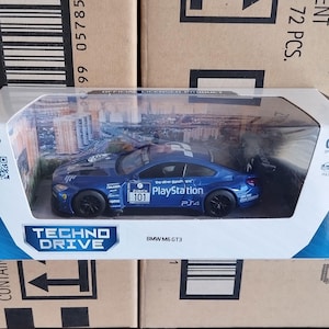 May include: A blue toy car model of a BMW M6 GT3 with the words "The other drive" and "PlayStation" on the side. The car is on a white display base with the words "Techno Drive" and "BMW M6 GT3".