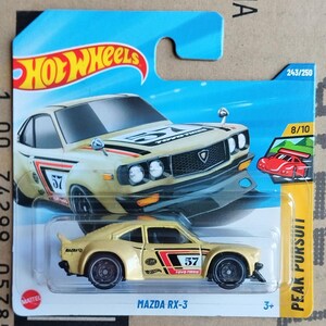 May include: A die-cast Mazda RX-3 toy car in a gold color scheme, featuring the number 57 on the sides and hood. The car has black and red accents, with the Hot Wheels logo visible. The packaging includes the text "Peak Pursuit".