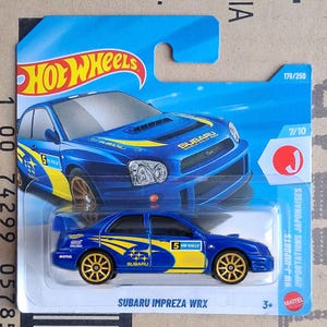 Könnte beinhalten: Ein blaues Hot Wheels Subaru Impreza WRX Modellauto in der Originalverpackung. Das Auto hat gelbe Seitengrafiken, goldene Räder und die Nummer 5. Die Verpackung enthält das Hot Wheels Logo, den Text Subaru Impreza WRX und die Angabe 176/250.