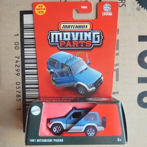 May include: A Matchbox Moving Parts die-cast model of a 1991 Mitsubishi Pajero SUV. The blue and white toy car is displayed in its original packaging, with the doors open. The packaging includes the Matchbox logo and the text "NEW NOUVEAU 2020".
