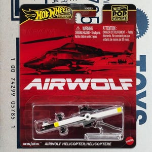 May include: Hot Wheels Premium Airwolf die-cast helicopter toy. The packaging is red with a graphic of the helicopter and the text "AIRWOLF". The toy helicopter has a white and black rotor with yellow accents. The packaging includes a warning about small parts.