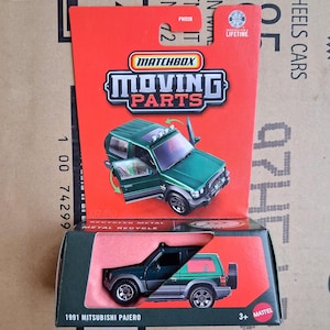May include: A Matchbox Moving Parts die-cast toy car of a 1991 Mitsubishi Pajero. The green SUV has opening doors and is packaged in a red and black box. The packaging includes the text "RECYCLED METAL" and "3+ MATTEL".