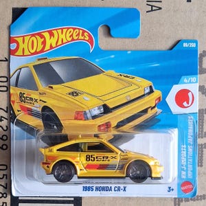 Op de afbeelding: Een gele 1985 Honda CR-X speelgoedauto van gegoten metaal. De auto heeft zwarte wielen en rode en zwarte accenten. De verpakking bevat het Hot Wheels-logo en de tekst "85 CR-X". De auto maakt deel uit van de HW J-Imports-serie.