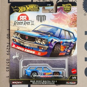 Op de afbeelding: Een Hot Wheels Premium modelauto, een blauwe Mazda RX-3 stationwagen. De auto heeft rode en blauwe accenten, met de tekst "TOYO TIRES" en "FURSTY". De verpakking bevat de tekst "RONIN RUN II" en "MAD MIKE MAZDA RX-3 WAGON/REMORQUE".