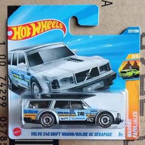 Könnte beinhalten: Ein silber-blaues Hot Wheels Volvo 240 Drift Wagon Spielzeugauto. Das Auto hat die Nummer 240 auf der Seite und ist auf einer blau-weißen Karte verpackt. Die Verpackung enthält das Hot Wheels-Logo und den Text "Wagons Familiales".