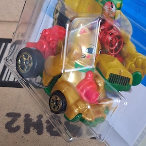 May include: A gold-colored toy car with a cat-like design, featuring a red engine and wheels with gold rims. The car is packaged in a clear plastic container. The car has a cartoonish design.