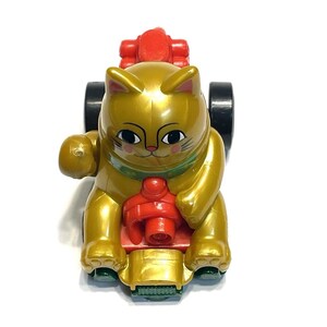 May include: A gold-colored toy cat with black wheels and red accents. The cat is in a seated position with one paw raised. The toy has a green collar and is designed to resemble a Maneki-neko, a Japanese lucky cat.
