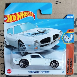 May include: A white '73 Pontiac Firebird die-cast model car on a blue and orange Hot Wheels card. The car features black wheels and a blue and black graphic on the hood. The card has the Hot Wheels logo and the text "Muscle Mania".