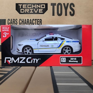 Op de afbeelding: Een witte 2015 Mustang politieauto speelgoed met blauwe en gele strepen, een rode lichtbalk en het woord "ПОЛІЦІЯ" aan de zijkant. Het speelgoed zit in een rood-witte doos met de tekst "TECHNO DRIVE TOYS".
