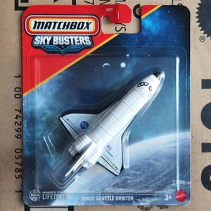 Op de afbeelding: Een Matchbox Sky Busters Space Shuttle Orbiter speelgoed, wit met zwarte en blauwe details, verpakt op een rode en blauwe kaart. Het speelgoed heeft het NASA-logo en het woord "Endeavour". De verpakking bevat de tekst "Designed for Lifetime".