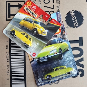 May include: Two Matchbox die-cast toy car models on their packaging. The first is a yellow 1970 Citroen 2CV with a black stripe on the side. The second is a yellow 1960 DS 21 Pallas with a black stripe on the side. Both cars are on their respective packaging with the Matchbox logo and the words "Lifetime" and "Mattel".