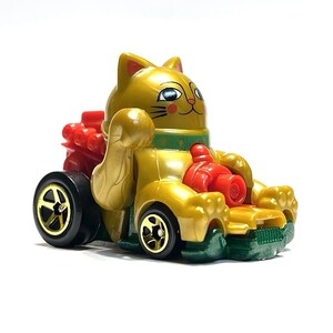 May include: A gold-colored toy car shaped like a Maneki-neko cat, with red and green accents. The cat is waving its right paw. The car has black wheels with gold rims. The toy is on a white background.