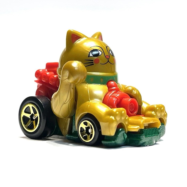 May include: A gold-colored toy car shaped like a Maneki-neko cat, with red and green accents. The cat is waving its right paw. The car has black wheels with gold rims. The toy is on a white background.