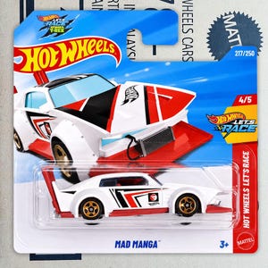 Może przedstawiać: Odlewany samochodzik Hot Wheels Mad Manga w kolorze białym z czerwonymi i czarnymi akcentami graficznymi, wyposażony w złote koła i wyraźny tylny spojler, zapakowany na niebieskiej karcie typu blister z napisem Hot Wheels Let's Race oraz 217/250.