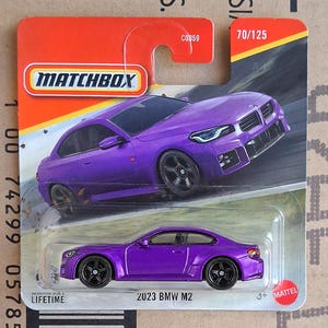 May include: A purple 2023 BMW M2 die-cast toy car in its original Matchbox blister packaging. The card features an illustration of the vehicle, the Matchbox logo, the text 70/125, and the label 2023 BMW M2 at the bottom of the plastic bubble.