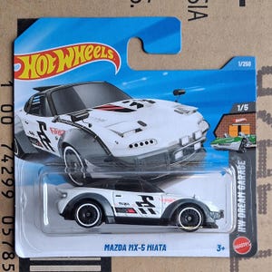 May include: A white and black Mazda MX-5 Miata die-cast toy car from the Hot Wheels HW Dream Garage series. The car features black wheels, a black roof, and grey accents. The packaging includes the Hot Wheels logo and the text "MAZDA MX-5 MIATA".