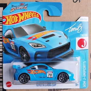 Może przedstawiać: Jasnoniebieski samochodzik Hot Wheels, Toyota GR86 Cup, na niebiesko-białej karcie. Samochód ma numer 70 i markę Hot Wheels. Opakowanie zawiera tekst "HW J-IMPORTS".