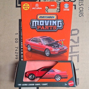 Può includere: Un'auto giocattolo Matchbox Moving Parts 1991 Acura Legend Coupe in confezione rossa. La carta presenta il logo Acura, il testo Matchbox Moving Parts e un'illustrazione dell'auto rossa con il cofano aperto. L'auto è visibile nella scatola.