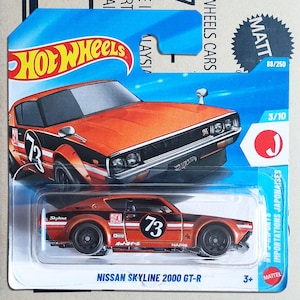 Könnte beinhalten: Orange-schwarzes Hot Wheels Nissan Skyline 2000 GT-R Spielzeugauto. Das Auto hat die Nummer 73 auf der Seite und einen weißen Streifen. Die Verpackung enthält das Hot Wheels-Logo und den Text "Importations Japonaises".